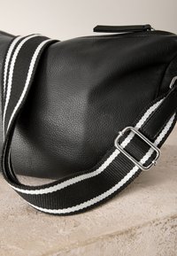 Sac en cuir noir texturé avec une bandoulière en tissu rayé noir et blanc réglable, posé sur une surface en pierre.