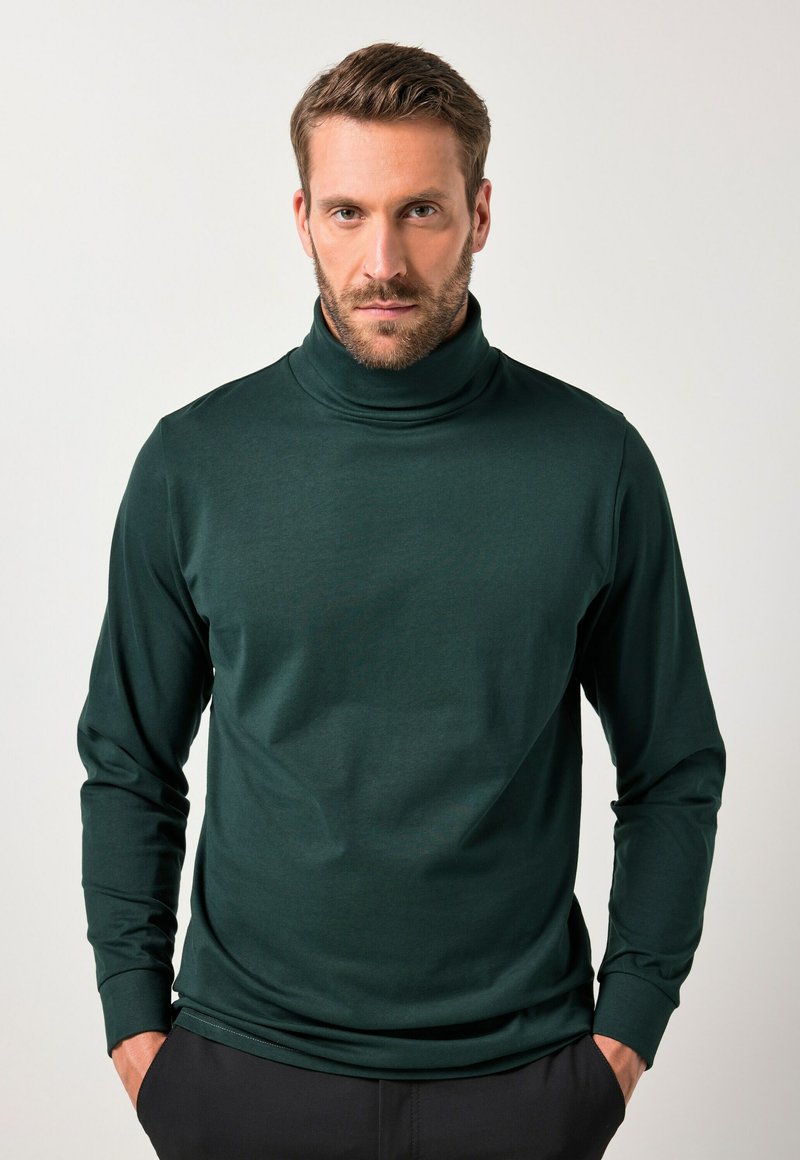 Tumma vihreä poolopaita, joka on valmistettu sileästä kankaasta. Paidassa on pitkät hihat ja se on muotoiltu istuvaksi. Kädet ovat taskuissa.