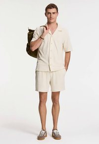 Camisa de manga corta beige claro y pantalones cortos a juego hechos de tela texturizada. El modelo lleva una mochila verde oscuro y usa zapatillas marrones y blancas.