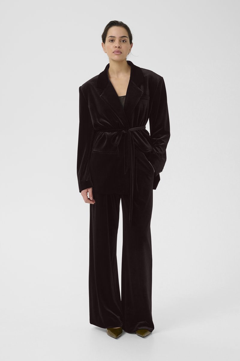 Schwarzer Samtblazer mit gebundenem Bund, breiten Ärmeln und einer Tasche, kombiniert mit passenden weit geschnittenen Hosen und spitzen grünen Schuhen.