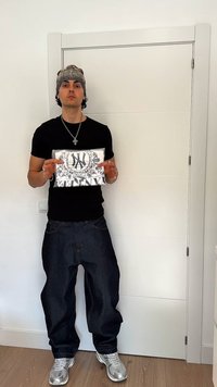 Νέος άνδρας με μαύρο μπλουζάκι, φαρδιά τζιν, γκρι σκούφο και ασημένια αθλητικά παπούτσια κρατάει μια μαύρη και λευκή τσάντα χειρός μπροστά από μια λευκή πόρτα.