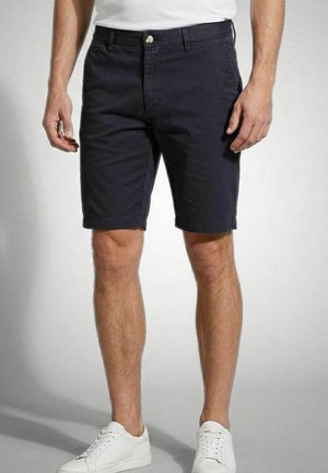 Man draagt zwarte knielange shorts, witte sneakers en een wit overhemd, staand tegen een effen achtergrond.