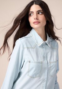 Light blue denim shirt featuring a pointed collar, two chest pockets, button front, and frayed collar detail. Smooth texture.