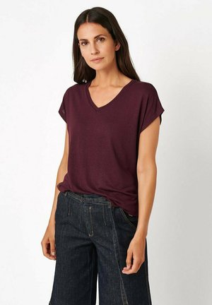 Frau mit langen dunklen Haaren trägt ein kurzärmeliges lila V-Ausschnitt-Shirt und dunkelblaue Jeans vor einem schlichten weißen Hintergrund.