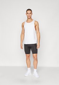 Hvid atletisk tanktop med brede stropper, parret med sorte tætsiddende shorts. Modellen har hvide sneakers på, og der er et logo på tanktoppen og shortsene.
