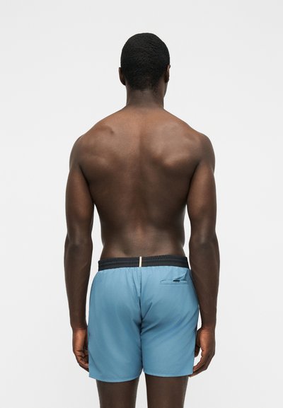Homme debout, dos face à l'avant, portant un short de bain bleu clair avec une ceinture noire et une poche arrière sur le côté droit.