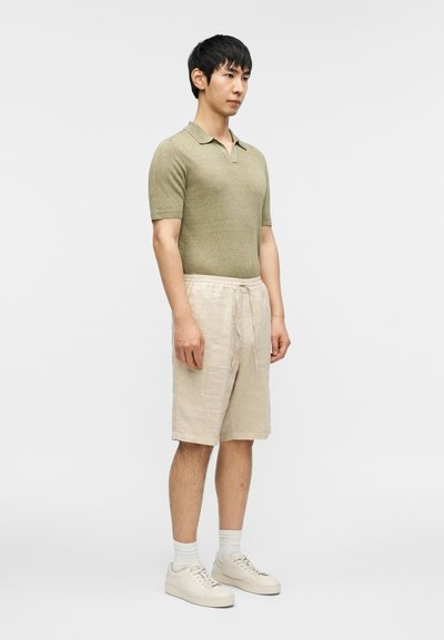 Φωτεινό πράσινο πόλο μπλουζάκι με κοντά μανίκια συνδυασμένο με μπεζ λινά σορτς. Το μοντέλο φοράει ανοιχτόχρωμα αθλητικά παπούτσια και κάλτσες, στέκεται μπροστά σε απλό φόντο.