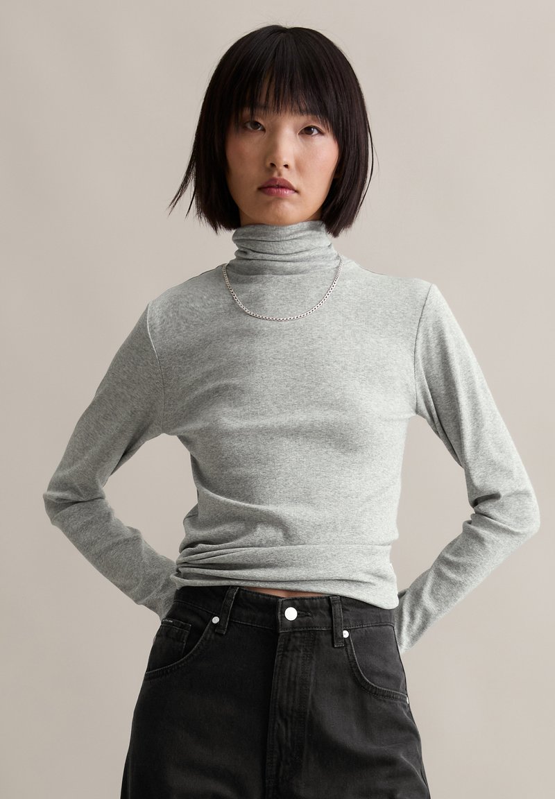 Lichtgrijze langslijf turtleneck van zacht, geribbeld stof met een strakke pasvorm. Gecombineerd met hoge zwarte denim. Zilveren ketting.