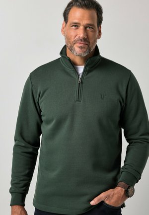 Mand med kort mørkt hår og skæg iført en mørkegrøn quarter-zip trøje og et armbåndsur, stående foran en ensfarvet lys baggrund.