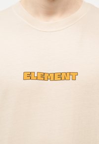 Μπεζ κοντομάνικο μπλουζάκι με στρογγυλή λαιμόκοψη και τη λέξη «ELEMENT» με έντονα, πορτοκαλί γράμματα σε μπλοκ, στο κέντρο του στήθους.