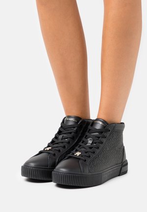 Baskets montantes noires avec des panneaux latéraux texturés, des lacets noirs et des accents de logo dorés portées sur des jambes nues sur un fond blanc.