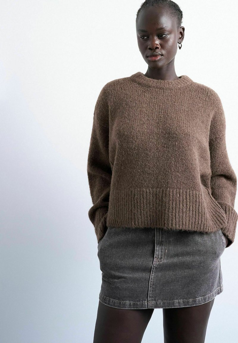 Femme portant un pull en maille marron avec les mains dans les poches et une jupe courte en denim noir délavé, sur un fond uni.