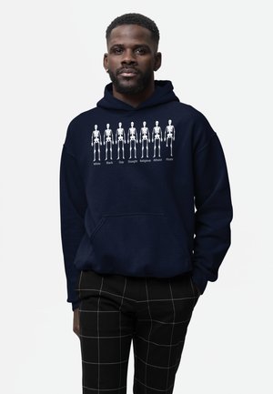 Námořnická mikina s bílými grafikami kostry označenými různými identitami. Měkká tkanina, volný střih a kapsa klokanka.