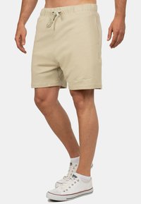 Pantaloni corti beige con vita elasticizzata e stringa, caratterizzati da una texture liscia e tasche laterali. Dettaglio del logo discreto sulla gamba.