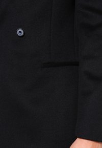 Cappotto nero in lana con chiusura a un bottone, revers eleganti e tasca laterale con una texture morbida.