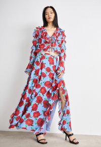 Floral two-piece outfit featuring a sky blue fabric with red roses, ruffle accents, long sleeves, and a high-slit skirt. Black strappy heels.