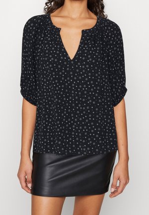 Frau trägt eine schwarze Bluse mit kleinen weißen Sternmustern und Dreiviertelärmeln, kombiniert mit einem schwarzen Minirock aus Leder.