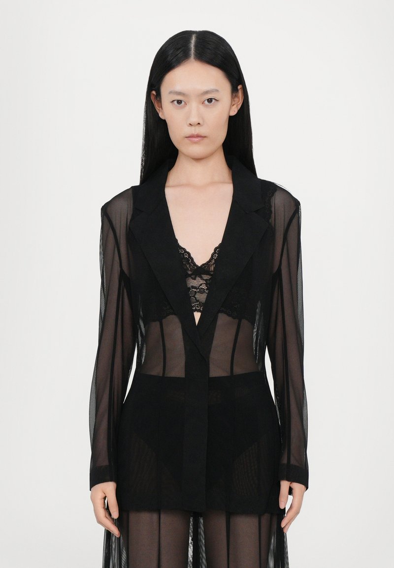 Durchsichtiger schwarzer Mantel mit langen Ärmeln und Reverskragen, über einem schwarzen Spitzen-Bralette getragen. Das Material wirkt leicht und transparent.