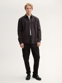 Camicia grigia scura con bottoni e tasca, maglietta bianca sotto, pantaloni neri e scarpe nere. L'aspetto complessivo è casual e aderente.