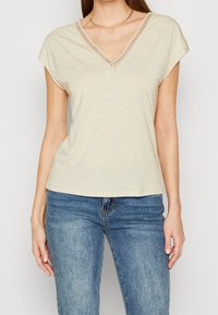 Beige V-hals t-shirt med korte ærmer, prydet med dekorative stikninger omkring halsudskæringen. Blødt stof med en afslappet pasform. Båret med blå jeans.