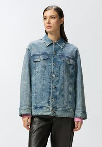 Traper jakna u svijetloplavoj boji s kopčanjem na gumbe, dva džepa na prsima i dekorativnim teksturiranim detaljima. Pretjerano širok kroj s manžetama.