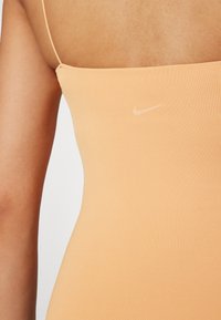 Nike'i ühtne bodi pehmest virsiku värvi kangast, mis omab siledat tekstuuri ja õhukesi rihmasid, väikese, heleda logoga prinditud õla lähedal.