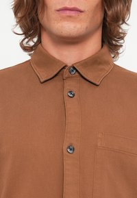 Bruine, gestructureerde blouse met een button-down kraag, voorzien van zwarte knopen en een borstzak, gepresenteerd tegen een witte achtergrond.
