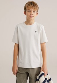 Bílé bavlněné tričko s kulatým výstřihem a krátkými rukávy, s malým černým logem na levé straně hrudi. Model drží pruhovaný svetr.