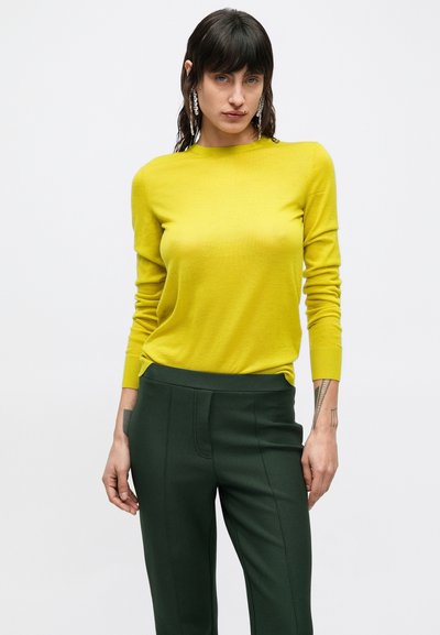 Geel nauwsluitend shirt met lange mouwen en een ronde hals, gecombineerd met donker groene, op maat gemaakte broek met verticale naden. Model poseert tegen een witte achtergrond.