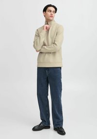 Beige halvzip genser i myk stoff, kombinert med løse blå denimjeans. Modellen står i en nøytral setting, og reflekterer et avslappet utseende.