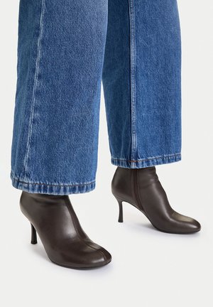 Ciemnobrązowe skórzane botki za kostkę z szpiczastymi noskami i wąskimi obcasami, noszone z szerokimi niebieskimi dżinsami z szerokimi nogawkami.
