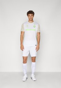 Biela futbalová dres s krátkym rukávom, so zelenými akcentmi a vertikálnymi pruhmi, spárovaný s bielymi šortkami a bielymi ponožkami po kolená.