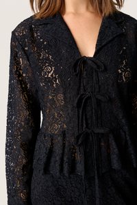 Must mustriga pluus kaelusega, V-kaelusega ja esise side detailiga. Kanga pinnal on keerukad lillemustrid ja kihiline peplum alläär.