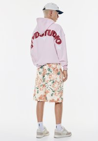 Roze hoodie met de tekst "STUDIO" in kastanjebruin, gecombineerd met knielange shorts met bloemenpatroon in crème, oranje en groen, en sneakers.