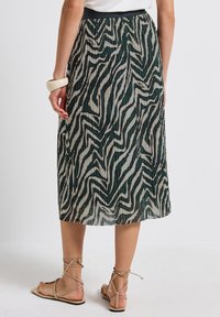 Pleated midi-hame, jossa on vihreä ja kerma-värinen zebra-kuvio, musta vyötärö ja sileä rakenne. Yhdistetty beigeihin sandaaleihin.