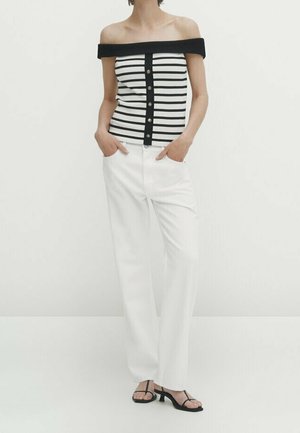 Donna che indossa un top a righe bianche e nere con bottoni e spalle scoperte, pantaloni bianchi a gamba dritta e sandali neri con tacco e punta aperta, mani nelle tasche.