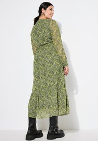 Abito lungo verde floreale con maniche lunghe arricciate, gonna a strati e sovrapposizione semi-trasparente. Abbinato a robusti stivali neri.