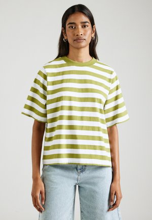 Vrouw met lang donker haar die een groen-wit horizontaal gestreept T-shirt met korte mouwen en lichtblauwe spijkerbroek draagt, staand tegen een effen achtergrond.