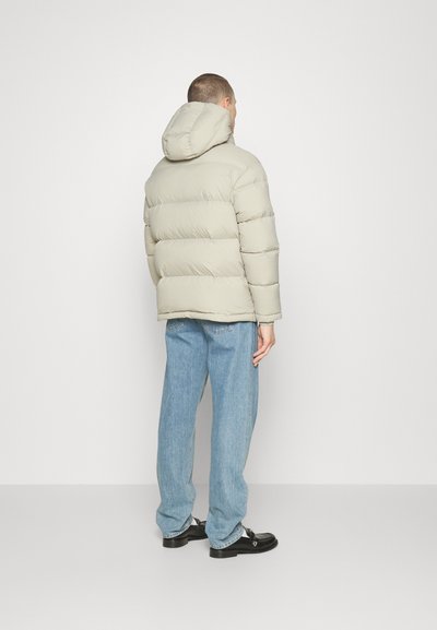 Pufferjakke i lysegrå med horisontale kviltede mønster og hætte, sat sammen med løstsiddende blå jeans og sorte sko.