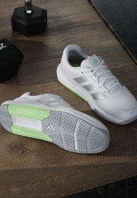 Paire de chaussures de sport blanches avec des accents vert clair sur la semelle, posée sur un sol en bois près de poids noirs.