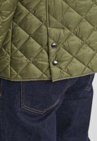 Olivgrüne, gestepfte Jacke mit Druckknöpfen am Saum, kombiniert mit dunkelblauen Jeans aus Denim mit sichtbaren Nähten und Taschen.