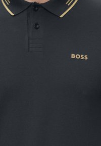 Čierne polo tričko s dvoma gombíkmi, zlatými pruhmi na okrajoch goliera a zlatým logom "BOSS" na ľavej strane hrudníka.