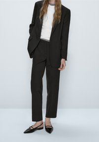 Czarny garnitur szyty na miarę, składający się z dopasowanej marynarki i prostych spodni, połączony z białą bluzką oraz czarnymi, szpiczastymi butami na odkrytej pięcie.