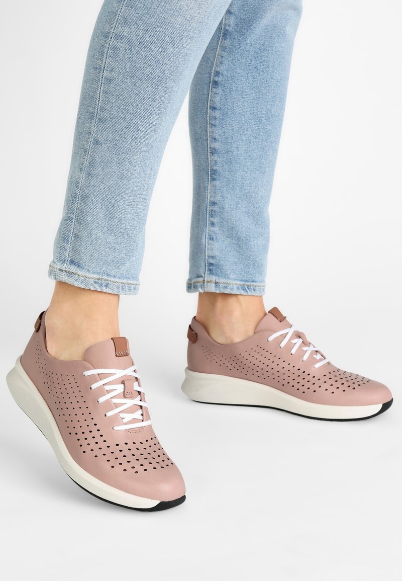 Pieds portant des baskets perforées rose clair avec des lacets blancs et des semelles blanches épaisses, associées à un jean bleu clair retroussé.