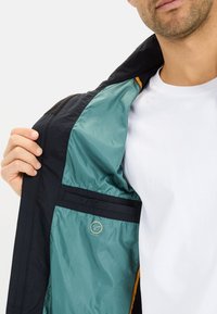 Czarna kurtka z turkusowym podbiciem, z logo, detalem zamka błyskawicznego i pomarańczowymi akcentami na kołnierzu. Gładka tekstura, lekka konstrukcja.