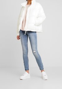 Chaqueta de plumas blanca con un collar alto, puños acanalados y cierre de cremallera, combinada con un suéter de punto rosa claro y jeans azul desgastados.