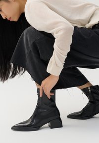 Bottines en cuir noir avec un bout pointu, un laçage à l'avant et un talon carré. Le design comprend une fermeture éclair sur le côté intérieur et une texture lisse.