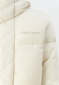 Krémově zbarvená prošívaná bunda s kapucí, se stehovanou texturou a viditelným šedým logem Tommy Jeans na hrudi.