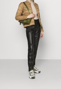 Person i tan paddad jacka, beige topp, svarta läderbyxor, gröna vita sneakers och grön axelväska mot en ljus enkel bakgrund.