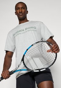 Sivý bavlnený tričko s nápisom "BJÖRN BORG TRAINING CLUB MONACO" potlačeným zelenou farbou. Čierne šortky. Drží čiernu a modrú tenisovú raketu s textúrovaným uchopením.